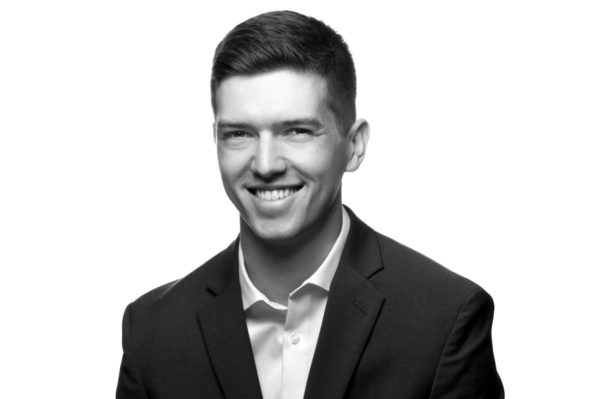 A professional black-and-white headshot features a young man with neatly styled short hair, wearing a dark suit jacket over a crisp white dress shirt. He is posed slightly at an angle while looking directly into the camera with a confident, approachable smile. The high-contrast lighting brings out the clean lines of his attire and the smooth contours of his face. Set against a pure white background, the composition is minimal and polished, conveying clarity and professionalism. This portrait is a refined representation of Business headshots Sonoma County.