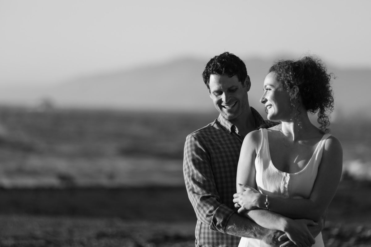 A smiling man in a checkered shirt embraces a woman with curly hair for a candid black and white couples photo. The joyful pair stands closely together at bodega bay with the woman wearing a white sleeveless top as natural light highlights their expressions against a blurred background of rolling hills.