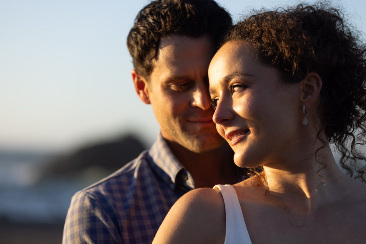 A smiling woman with curly brown hair in an updo wears a white top as she poses closely with her partner in a close up portrait of couple at sunset. The man with dark curly hair and a blue plaid shirt rests his head near hers with a gentle smile while warm golden sunlight illuminates their faces against the blurred ocean background.