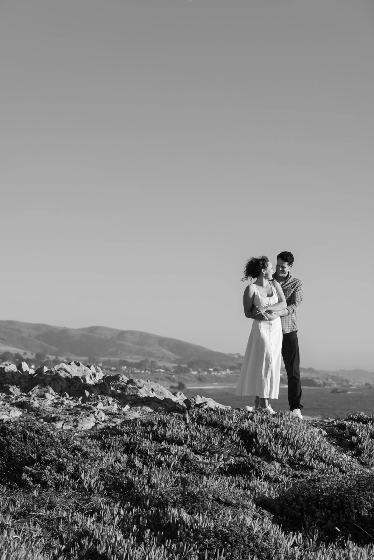 A woman with curly hair in a white sleeveless dress stands smiling up at a man wearing a checkered shirt as he embraces her on a rocky coastal bluff. The black and white photograph captures soft natural daylight across the ocean and distant hills in the background during their couples photography Sonoma County.
