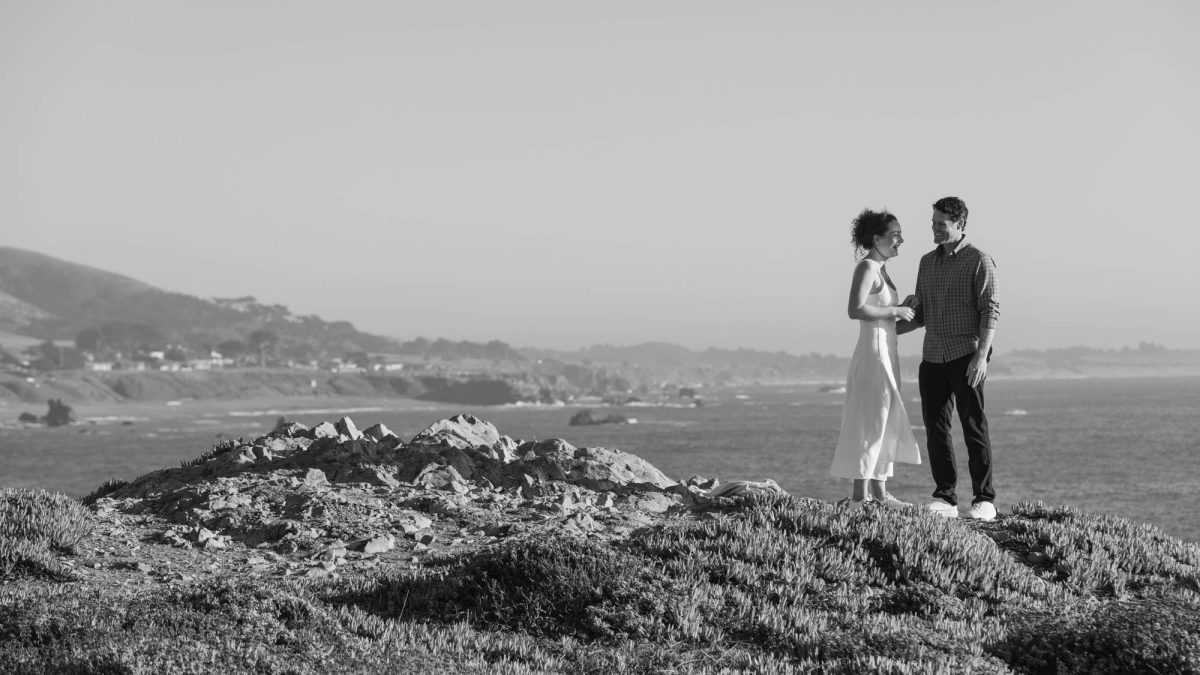 A woman with curly hair in a white sleeveless dress stands smiling at a man wearing a checkered shirt as they face each other on a rocky coastal bluff. The black and white photograph captures soft natural daylight across the vast ocean waves and distant hills in the background during their couples photos on the Sonoma County coastline.