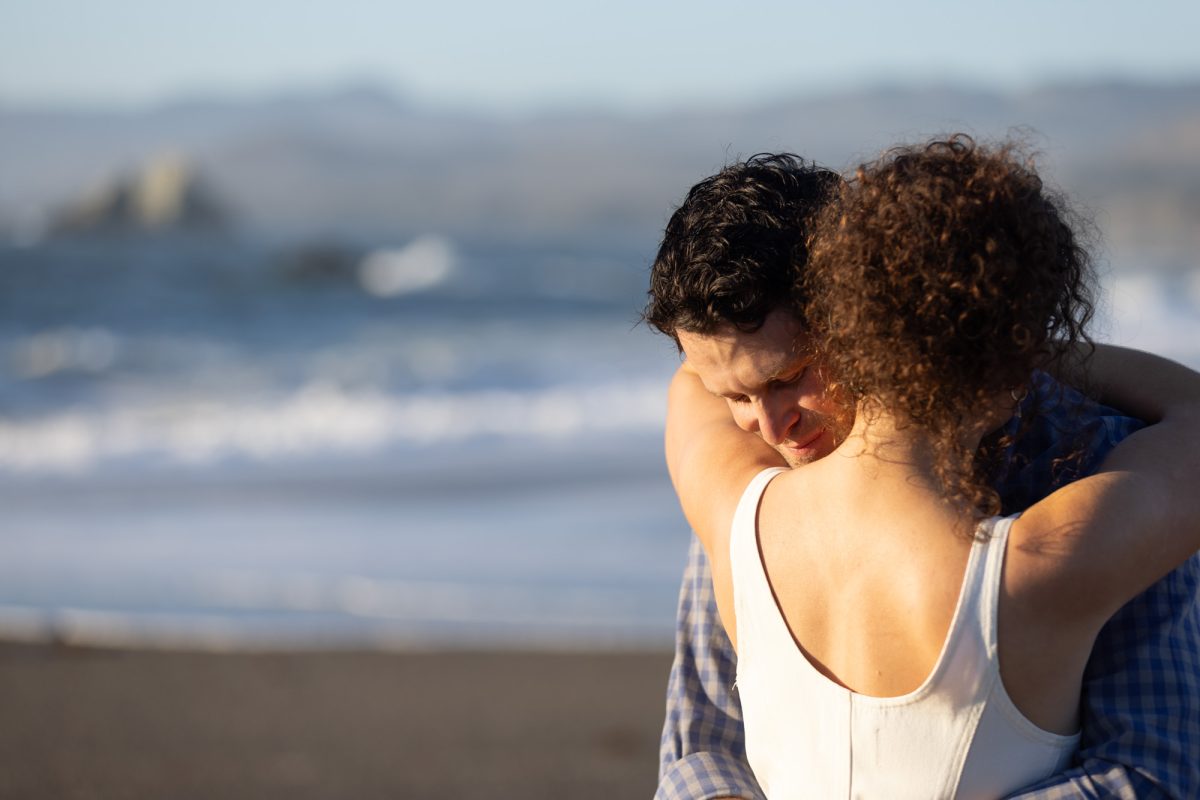 A woman with curly brown hair in a white top shares a tender embrace with her partner in an emotional couples portrait on the beach. The man with dark curly hair wearing a blue plaid shirt rests his head gently against her shoulder with a soft smile as warm golden sunlight illuminates their close moment against the blurred ocean waves rolling onto the sandy shore.