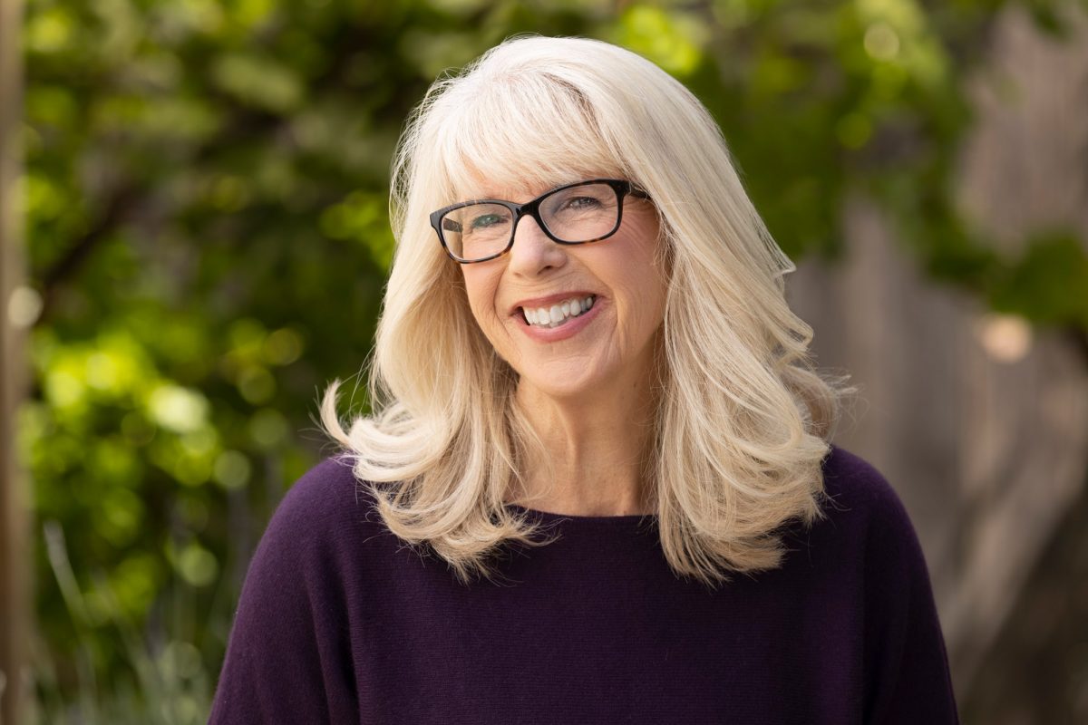 A woman with a warm, friendly expression stands in an outdoor setting with softly blurred greenery in the background. Her straight blonde hair is neatly styled in a chin-length bob, parted to the side. She wears a textured light pink blouse with a rounded neckline and subtle pattern, complemented by a single strand of pearls that adds a touch of classic elegance. Her bright smile and relaxed posture are accentuated by the gentle natural light that softly illuminates her features. This inviting and polished portrait is a fine example of Headshot photography Petaluma.