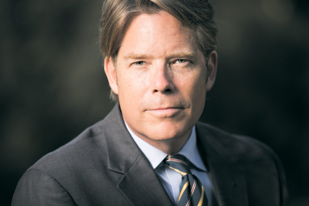 A professional man in a charcoal suit poses against a softly blurred dark background, captured in a close-up portrait with natural, directional lighting. His expression is composed and confident, with a direct gaze that conveys authority and focus. He wears a light blue dress shirt paired with a diagonally striped tie in gold, navy, and burgundy, knotted neatly at the collar. His medium-length hair is combed to the side, and the lighting highlights the natural texture of his skin and the sharp lines of his jaw. This image exemplifies the polished and trustworthy presence typical of Legal Headshots Sonoma County.