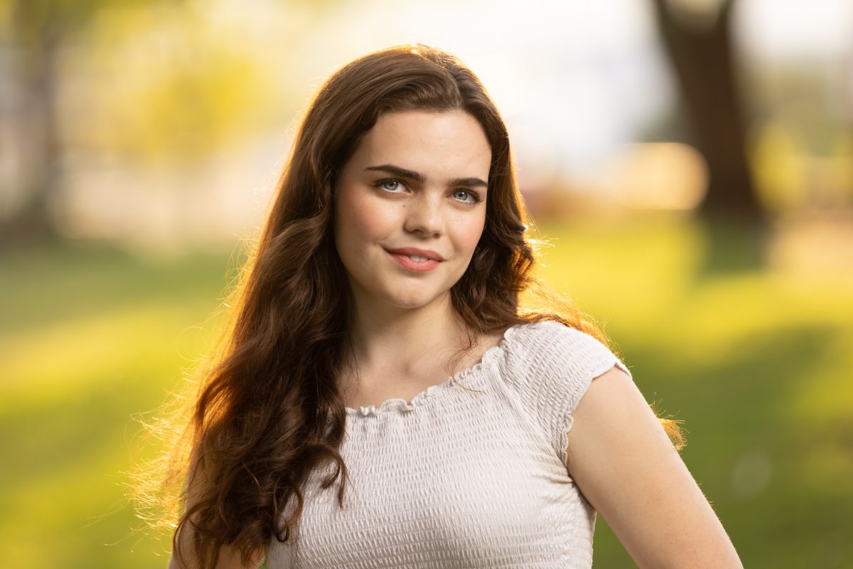 A young woman stands outdoors in golden-hour light, framed by a softly blurred background of green grass and glowing trees. Her long, wavy brown hair cascades over her shoulder, catching the warm sunlight at the edges. She wears a fitted, short-sleeved, smocked white top that complements her fresh and natural look. Her expression is poised and confident, with a gentle smile and direct eye contact. The lighting softly illuminates her features, highlighting her clear complexion and striking eyes. This portrait is a radiant example of Modeling Headshots Sonoma County.