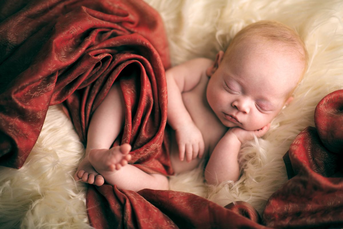 A peacefully sleeping newborn rests on a plush, ivory faux fur surface, creating a soft and serene setting. The baby's delicate skin has a warm, gentle glow, and their small hand is tucked under one cheek while the other arm lies loosely across their torso. A rich, red brocade fabric with subtle gold detailing is elegantly draped over and around the infant, partially covering their body and adding a touch of texture and warmth to the composition. The baby's tiny toes peek out from beneath the folds of the fabric, emphasizing their smallness and vulnerability. This tender and intimate moment is beautifully captured, highlighting the artistry of Newborn photography Sonoma.