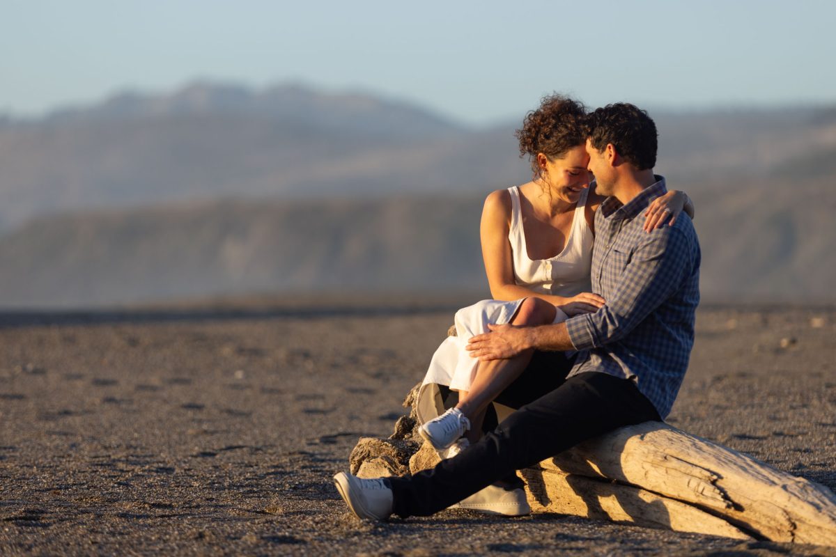 A woman with curly brown hair in a white top sits closely with her partner on a large driftwood log during their Northern California coastal couples portraits. The man with dark curly hair wearing a blue plaid shirt has his arm around her as they rest their foreheads together with smiles while warm golden sunlight illuminates their close moment on the sandy beach.