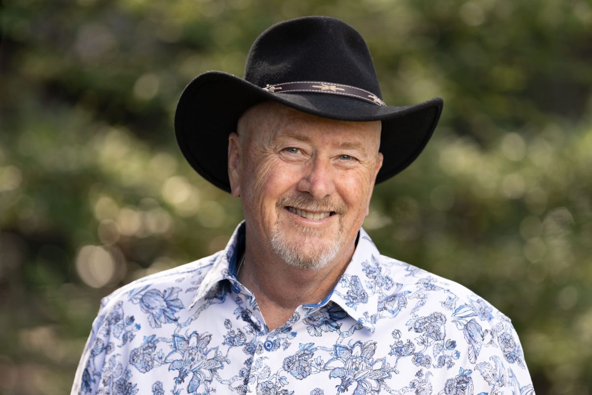 Headshot of Woody from "Woody's Wine Tours" stands outdoors, framed by a softly blurred background of green foliage and dappled sunlight. He wears a wide-brimmed black cowboy hat adorned with a decorative band, and a light-colored button-up shirt featuring a detailed blue floral print. His neatly groomed white goatee and warm smile convey a relaxed and approachable demeanor. The natural lighting and shallow depth of field focus attention on his face, enhancing the clarity of his features. This image captures the essence of Personal Branding Headshots Santa Rosa, blending personality and professionalism in a natural setting.