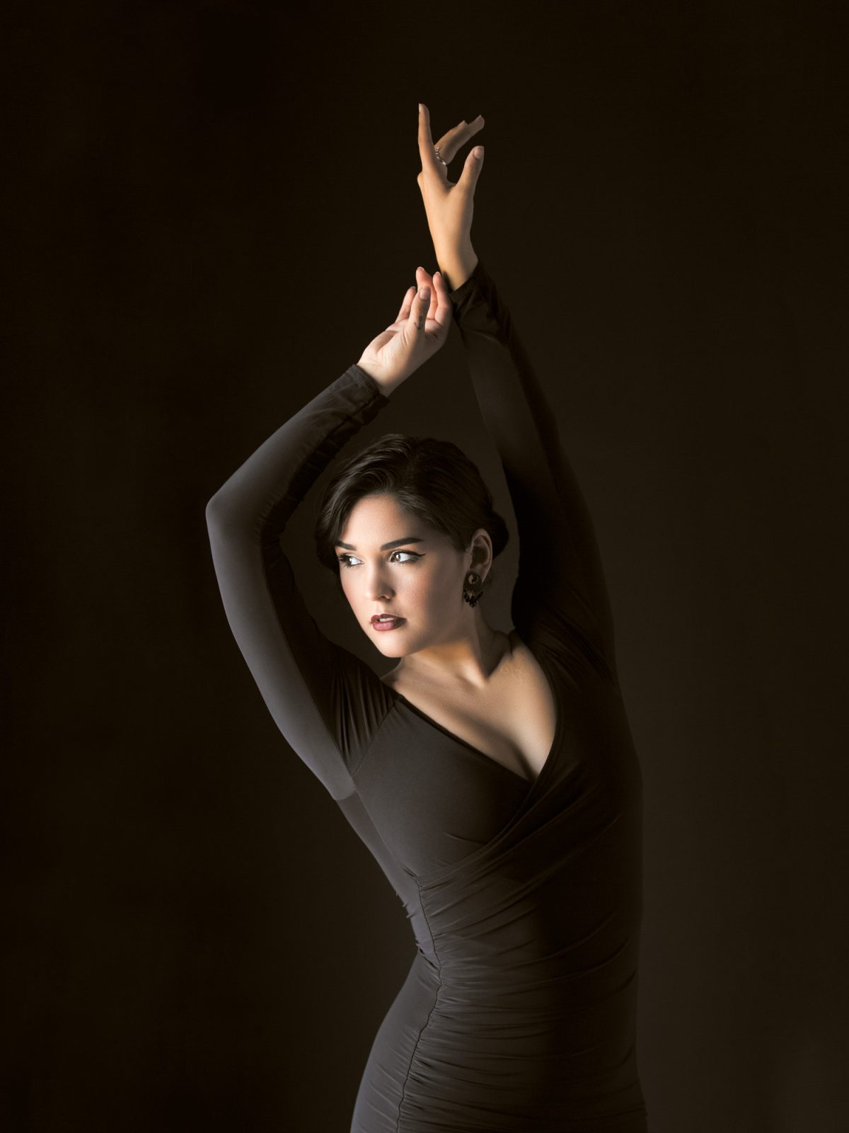 A striking studio portrait showcases a woman posed gracefully against a deep, dark background. She wears a form-fitting, long-sleeved black dress with a V-neckline and subtle ruching along the torso, which enhances the elegant lines of her figure. Her arms are extended upward in a fluid, ballet-like gesture, fingers delicately poised, creating a sense of movement and sophistication. Her dark hair is styled in a sleek, side-parted bob, and she wears bold makeup with rich red lipstick and defined eyeliner. Soft, directional lighting highlights the contours of her face and body while casting gentle shadows, lending depth and drama to the composition. This refined image exemplifies the artistry of Portrait photography Sonoma, California.