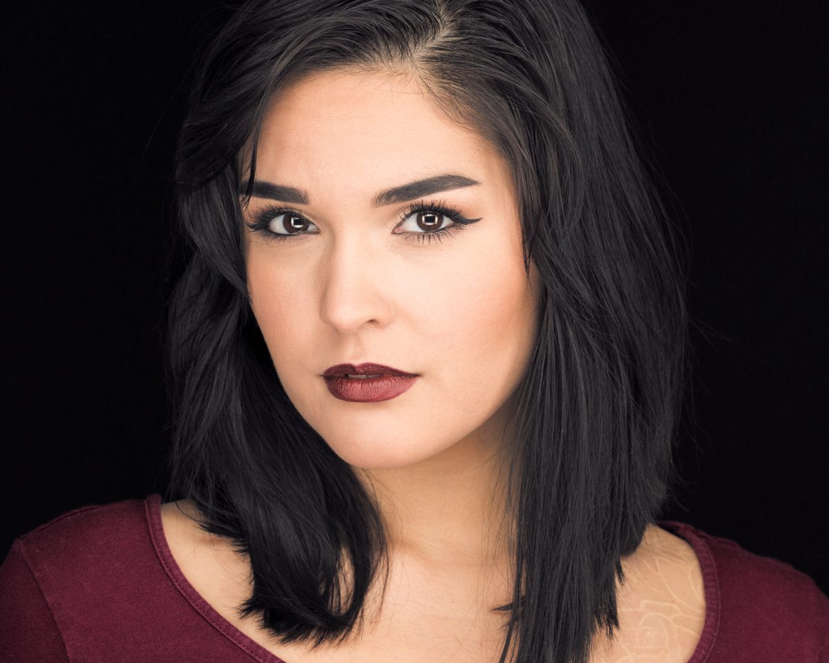 A woman poses against a seamless black background in a striking, high-contrast portrait. Her dark, shoulder-length hair frames her face in loose layers, with a side part adding definition to her style. She wears bold makeup, featuring sharply defined brows, dark winged eyeliner, and deep burgundy lipstick that complements her rich maroon top. Her expression is serious yet composed, with a confident gaze directed slightly off-camera. The lighting is soft and even, emphasizing the clarity and tone of her skin while casting subtle shadows that enhance depth. This refined portrait is an elegant example of work by a Profile photo photographer Sonoma County.