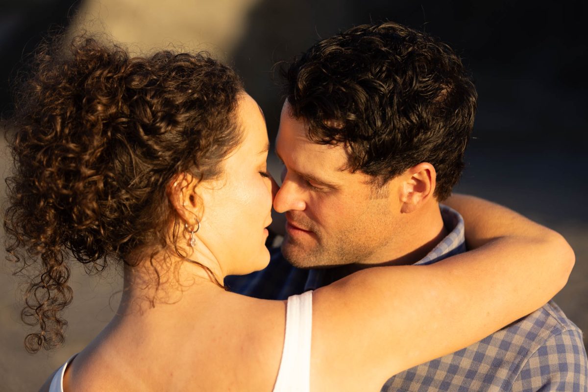 A woman with curly brown hair in an updo wearing a white top embraces her partner in a romantic candid couple portrait on the beach. The man with dark curly hair in a blue plaid shirt leans in close with their foreheads touching as warm golden sunlight illuminates their faces in a tender moment.