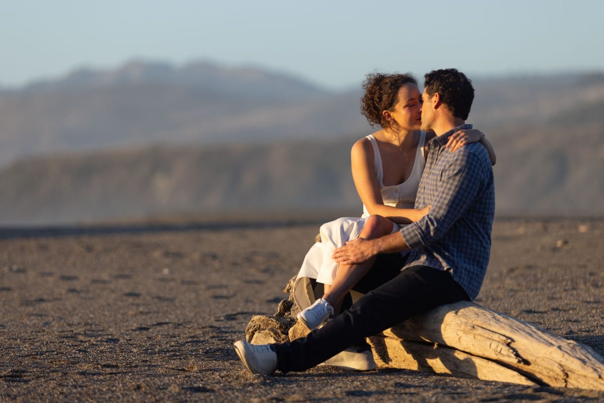 A woman with curly brown hair in a white top sits closely with her partner on a large driftwood log during romantic couple photos captured at Duncan's Landing. The man with dark curly hair wearing a blue plaid shirt has his arm around her as they lean in for a kiss while warm golden sunlight illuminates their close moment on the sandy beach.