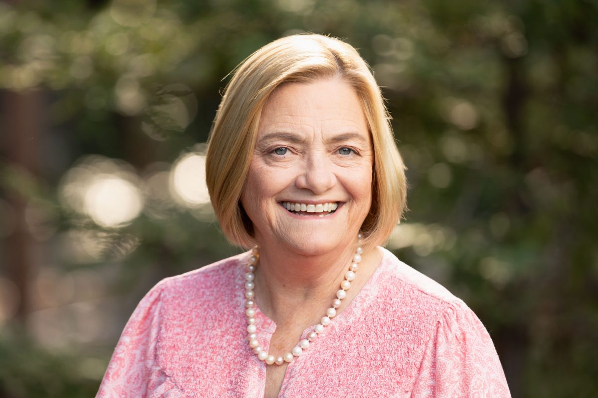 A smiling woman poses outdoors with a background of softly blurred green foliage and gentle sunlight filtering through the trees. She has short, straight blonde hair parted to the side, and her expression radiates warmth and approachability. She wears a textured pink blouse with a subtle paisley pattern and a classic string of pearls around her neck, adding a touch of elegance to her ensemble. The natural light illuminates her face evenly, enhancing her eyes and friendly demeanor. This image exemplifies the inviting and professional style characteristic of Santa Rosa Headshots.
