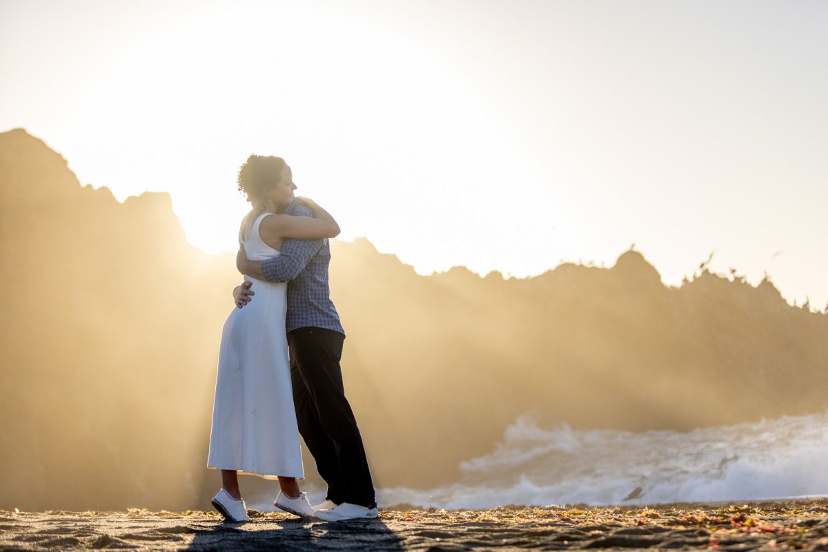 A dramatic golden-hour photograph from Sonoma County beach engagement sessions, portraying a tender embrace between a couple on a rugged coastal edge as the sun dips low, casting warm rays and ethereal haze over jagged rock formations and crashing waves below. The woman in a simple white dress leans into her partner in a checkered shirt, their silhouettes framed against the misty horizon, symbolizing enduring love amidst the wild beauty of the Pacific shore.