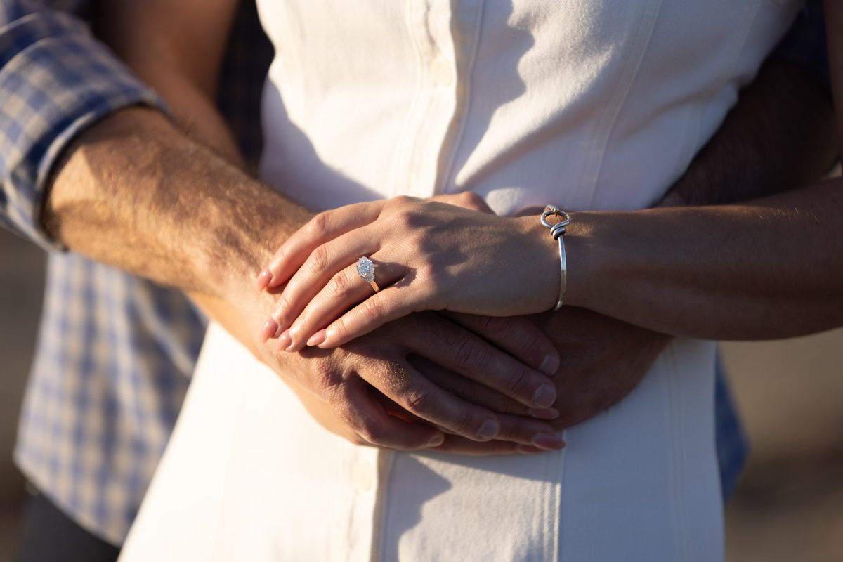 Close-up of a couple's intertwined hands during an engagement session by a Sonoma County Engagement Photographer, showcasing a brilliant oval-cut diamond ring on the woman's finger alongside a silver bracelet, her manicured nails and his strong arm embracing her white dress-clad waist, with soft warm sunlight casting gentle shadows, symbolizing love, commitment, and intimate connection against a blurred coastal backdrop.