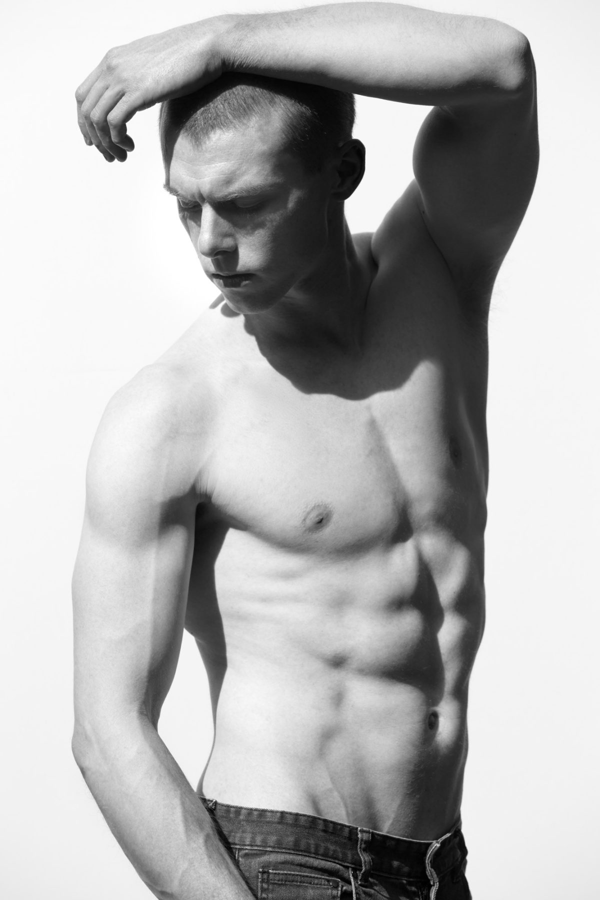 In this high-contrast black and white image, a young man poses shirtless against a plain, bright backdrop, highlighting his defined physique. His head is turned slightly downward and to the side, eyes closed, with one arm bent over his head, casting dramatic shadows across his chiseled face and upper body. The lighting emphasizes the muscular contours of his arms, chest, and torso, creating a sculptural effect. He wears dark jeans that rest just below his waist, completing the minimal and focused aesthetic. The composition captures strength, form, and precision, serving as a striking example of Sonoma County Fitness photography.