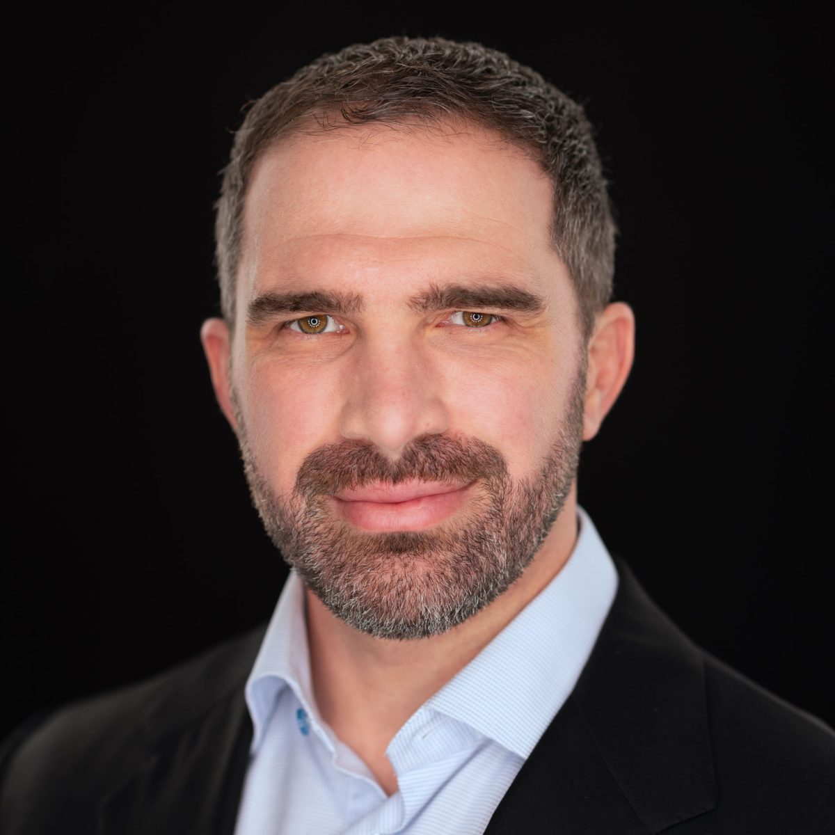 A man with short, neatly trimmed dark hair and a full, well-groomed beard poses confidently against a seamless black background. His expression is calm and self-assured, with a slight, professional smile and direct eye contact. He wears a light blue dress shirt under a black blazer, giving him a polished and business-ready appearance. The lighting is soft and even, bringing out the details in his eyes and facial features while maintaining a clean, professional finish. This image exemplifies the precise and refined style of Sonoma County Headshot Photographer Jason Guy.