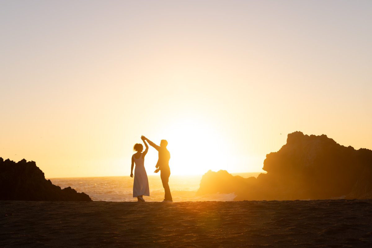 A woman with curly brown hair in a white dress holds her partner's hand high as they dance together during their Sonoma County proposal at Bodega Bay. The man with dark curly hair faces her while warm golden sunlight creates a dramatic silhouette of their moment on the sandy beach with the ocean and large rock formations behind them.