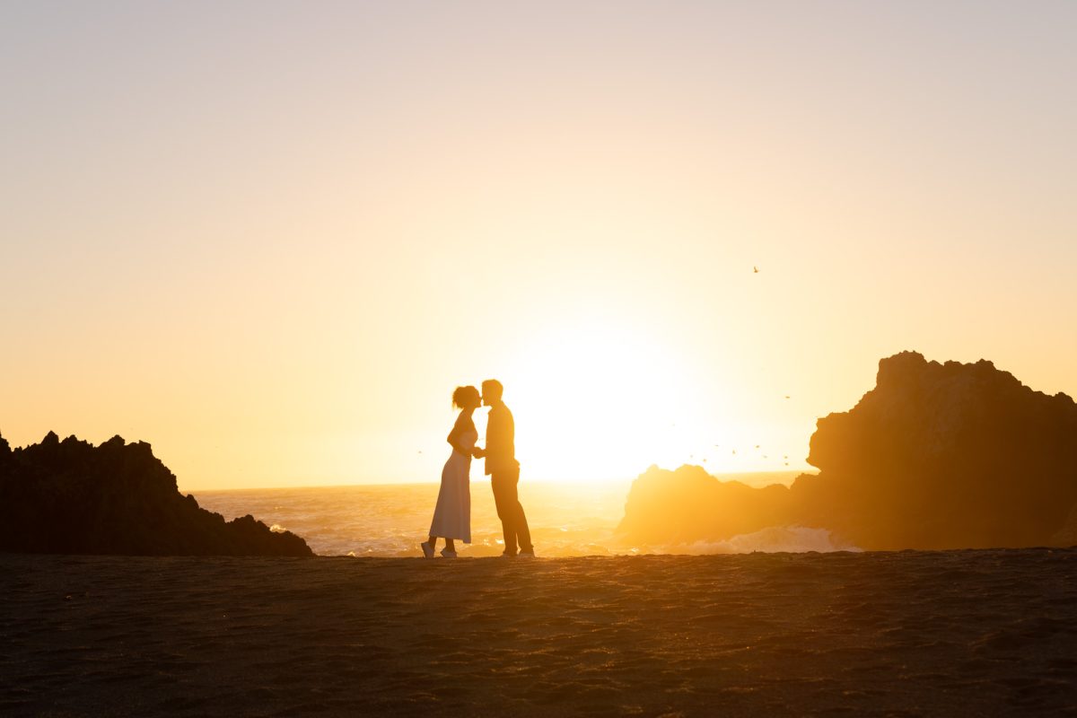 Silhouette of a romantic couple—a woman in a flowing white dress and a man in dark pants and shirt—sharing a tender kiss while standing on a sandy beach at the edge of the ocean, framed by jagged rocky cliffs on either side. The setting sun casts a warm golden glow across the scene, with gentle waves lapping at the shore and a few birds soaring in the gradient sky transitioning from deep orange to soft blue. Captured by Sonoma County sunset beach photographer.