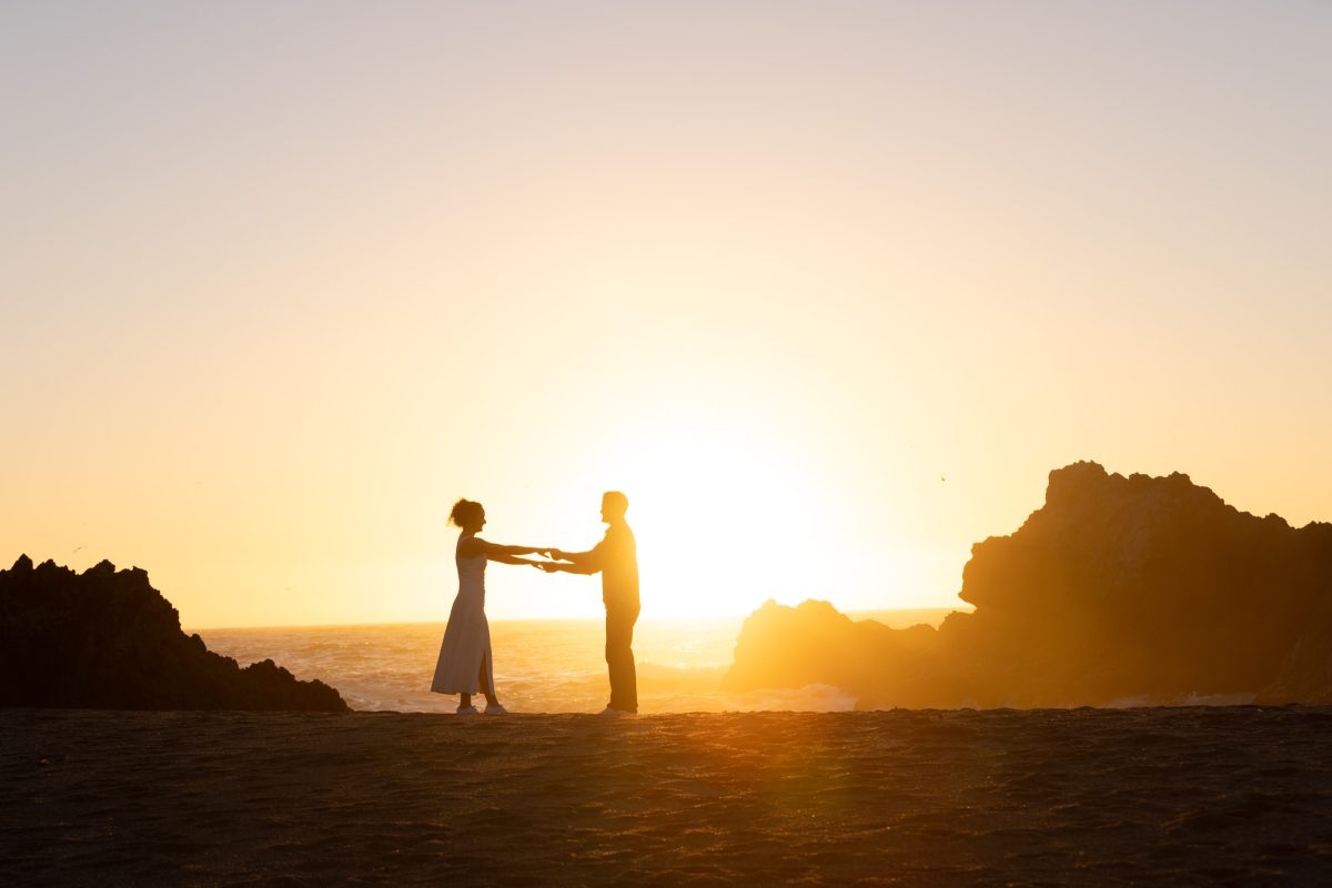 A woman with curly brown hair in a white dress holds hands with her partner during a surprise proposal at sunset. The man with dark curly hair stands facing her as warm golden sunlight creates a dramatic silhouette of their close moment on the sandy beach with the ocean and large rock formations behind them.