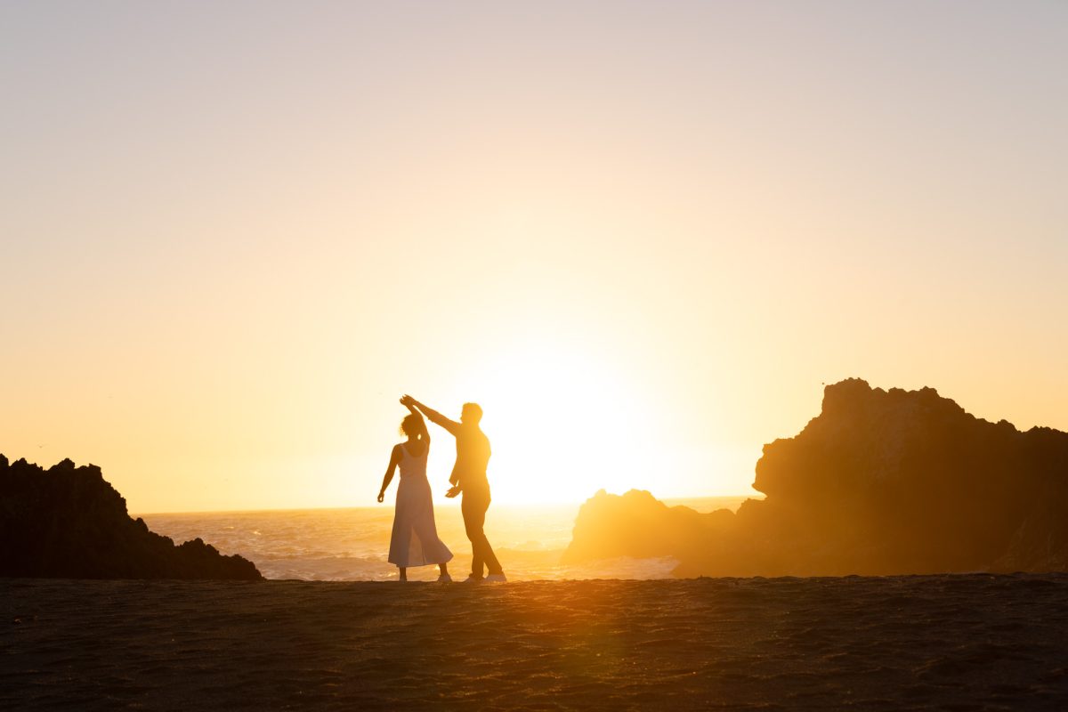 A woman with curly brown hair in a white dress holds her partner's hand high as they stand together during a Surprise Proposal in Bodega Bay, CA. The man with dark curly hair faces her while warm golden sunlight creates a dramatic silhouette of moment of them dancing on the sandy beach with the ocean and large rock formations behind them.