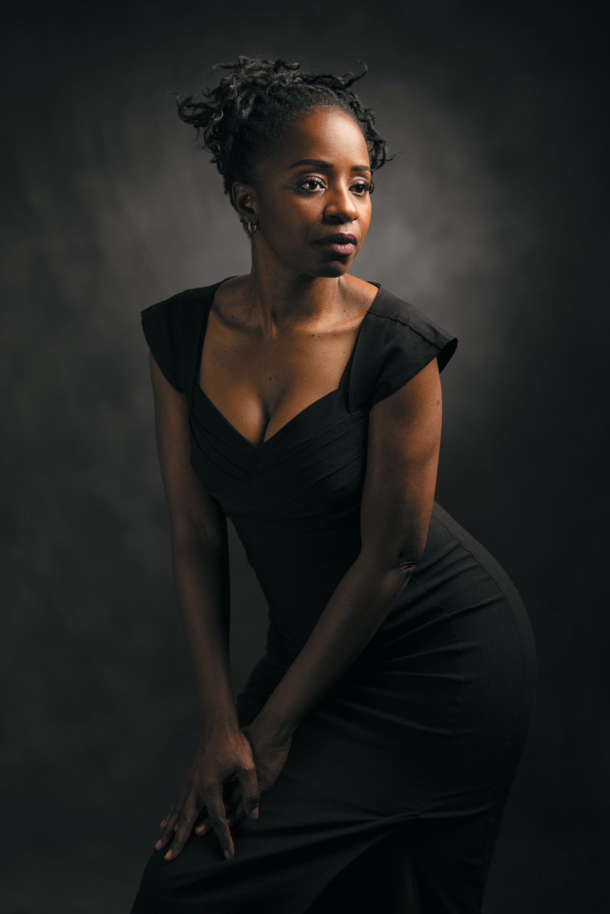 In this refined studio portrait, a woman poses gracefully against a softly textured dark background, illuminated by dramatic, directional lighting that accentuates the contours of her face and figure. She wears a fitted black dress with cap sleeves and a structured neckline, conveying elegance and strength. Her posture is poised and intentional, with both hands resting gently on her bent knee as she leans slightly forward, gazing contemplatively into the distance. Her natural hair is styled in an updo, and subtle gold earrings add a delicate touch to the composition. The image exemplifies the timeless elegance and emotional depth characteristic of Traditional portrait photography Sonoma.