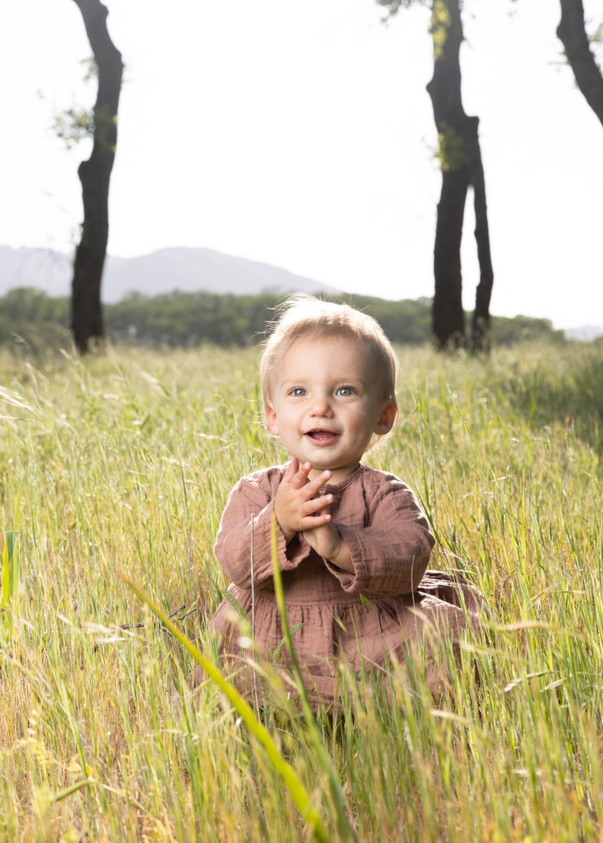 Young child with blonde hair smiles from seated position amid tall grass for Sonoma County kids portrait session. Bright light bathes green meadow in Sonoma County as her small hands clap together near a background trees.