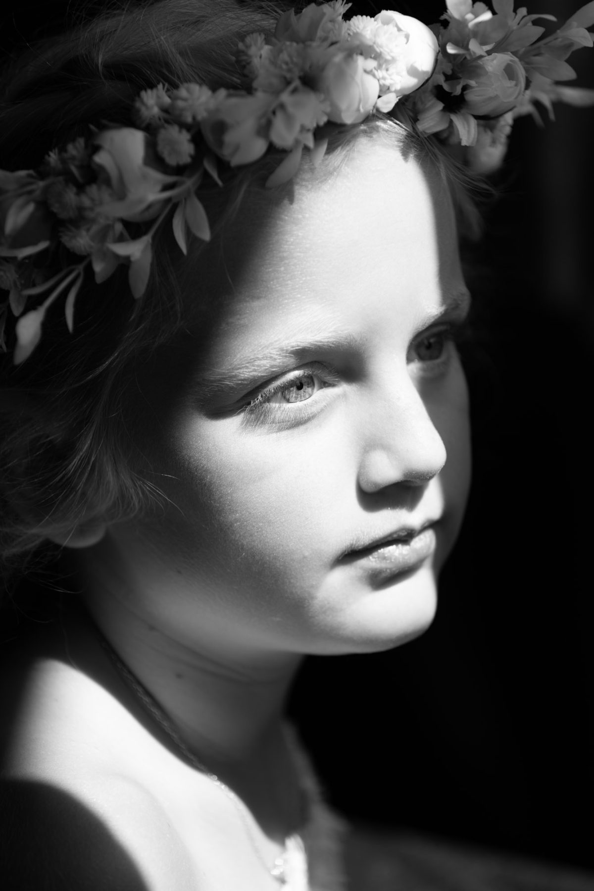 Dramatic lighting sculpts the gentle face of a young girl adorned with an intricate floral crown captured in artistic children's portrait session. The black and white image shows her looking peacefully away with a serene expression and soft shadows defining her profile.