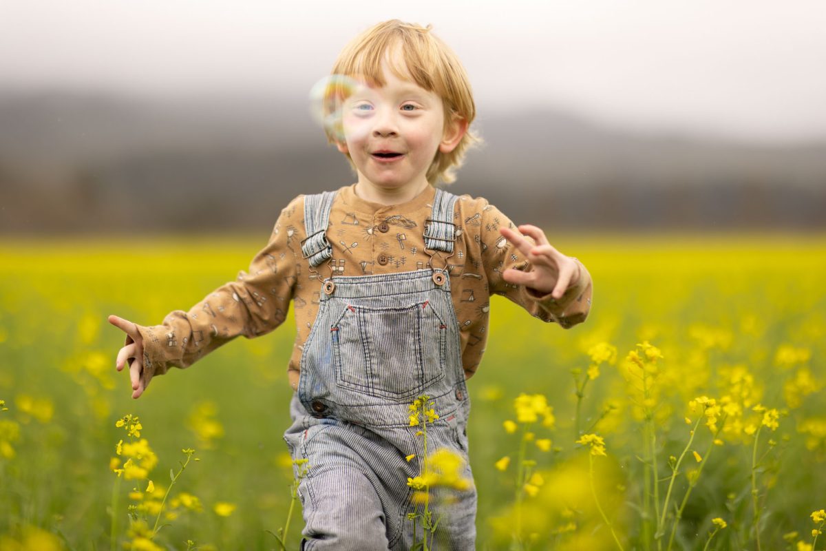 A joyful young boy with blond hair and blue eyes smiles brightly as he reaches out with both arms to chase a bubble through a vibrant field of yellow flowers. Soft natural light illuminates his patterned shirt and striped overalls in this playful outdoor scene.