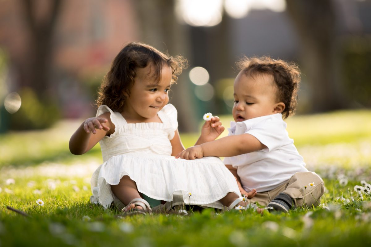 An older sister and her baby brother enjoy an authentic happy moment sitting on the grass surrounded by small daisies for their brother sister portrait. Soft sunlight highlights her white ruffled dress and his white shirt as they platy together. She holds a white daisy in her hand as her brother points to another laying on her dress. 