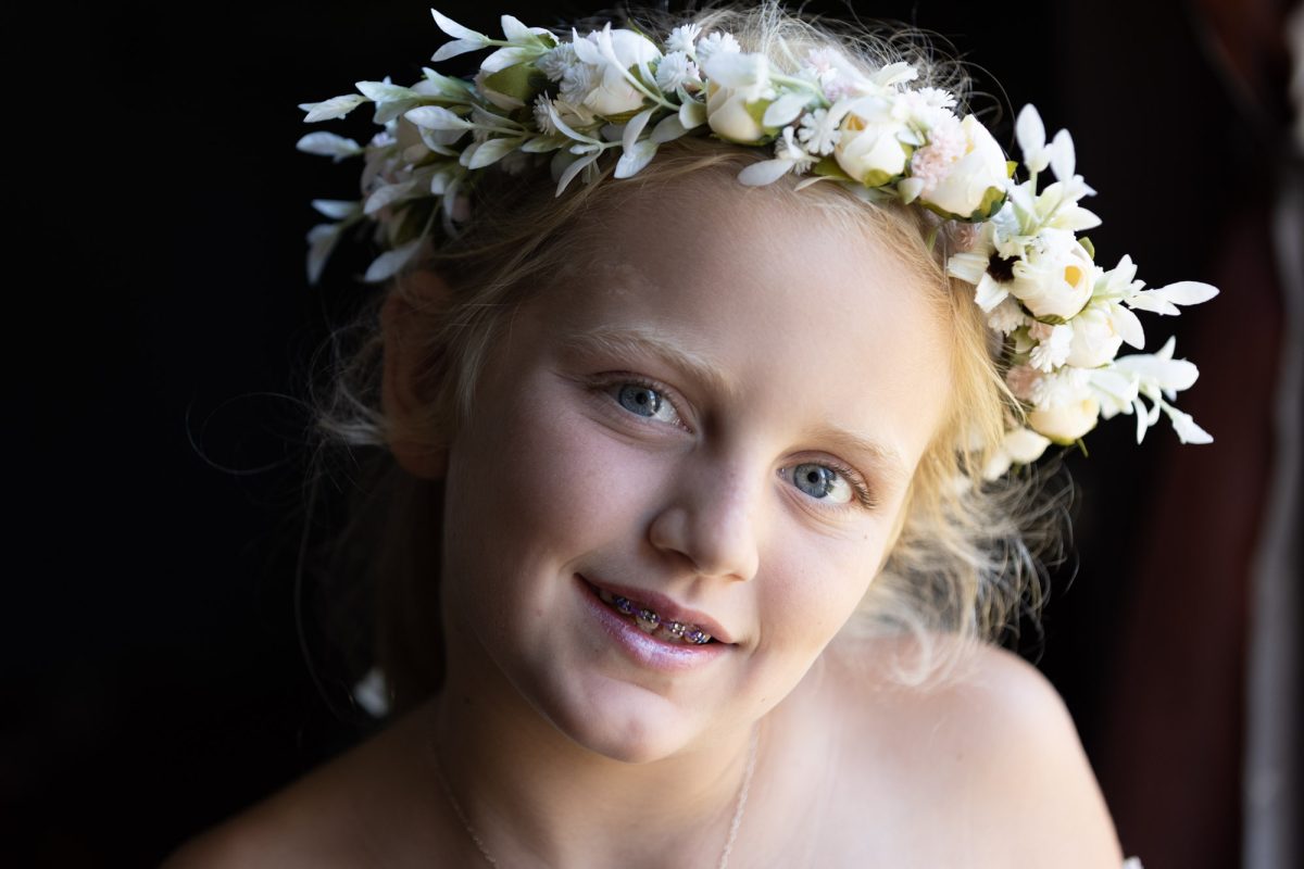 Soft lighting highlights the face of a young girl adorned with a delicate flower crown captured in this children's portrait session captured in Sonoma County. Her bright blue eyes shine as she smiles gently showing her colorful braces against a dark background.