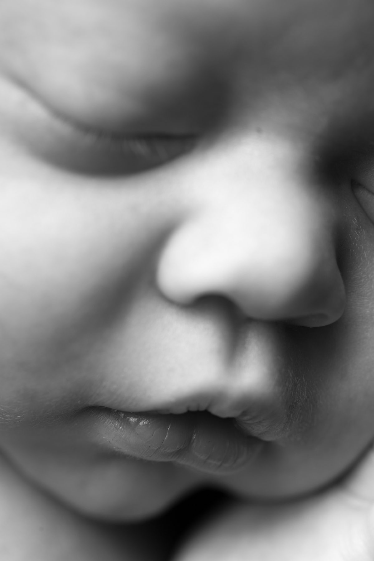 A sleeping baby with eyes gently closed is shown in this close up newborn portrait. Soft lighting creates gentle shadows that highlight the delicate nose, tiny mouth and smooth skin in soft black and white tones.
