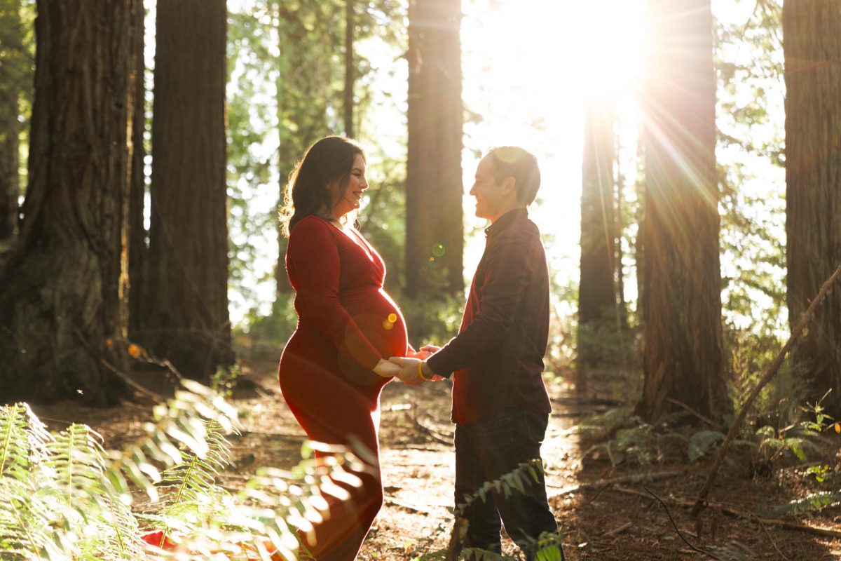 A pregnant woman in a long red dress and her husband smile at each other during their couples maternity photos in a redwood forest. They stand face to face holding hands over her belly as bright golden sunlight streams through the tall trees creating beautiful lens flare.