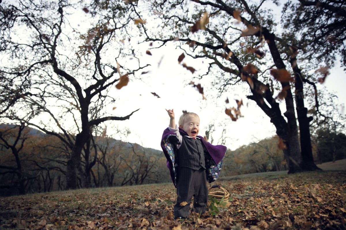 A joyful young boy throws handfuls of autumn leaves high into the air with an excited smile during his fall children's portraits in Sonoma County. Warm natural light illuminates his purple cape and pinstripe suit as he stands among fallen leaves beneath large oak trees.