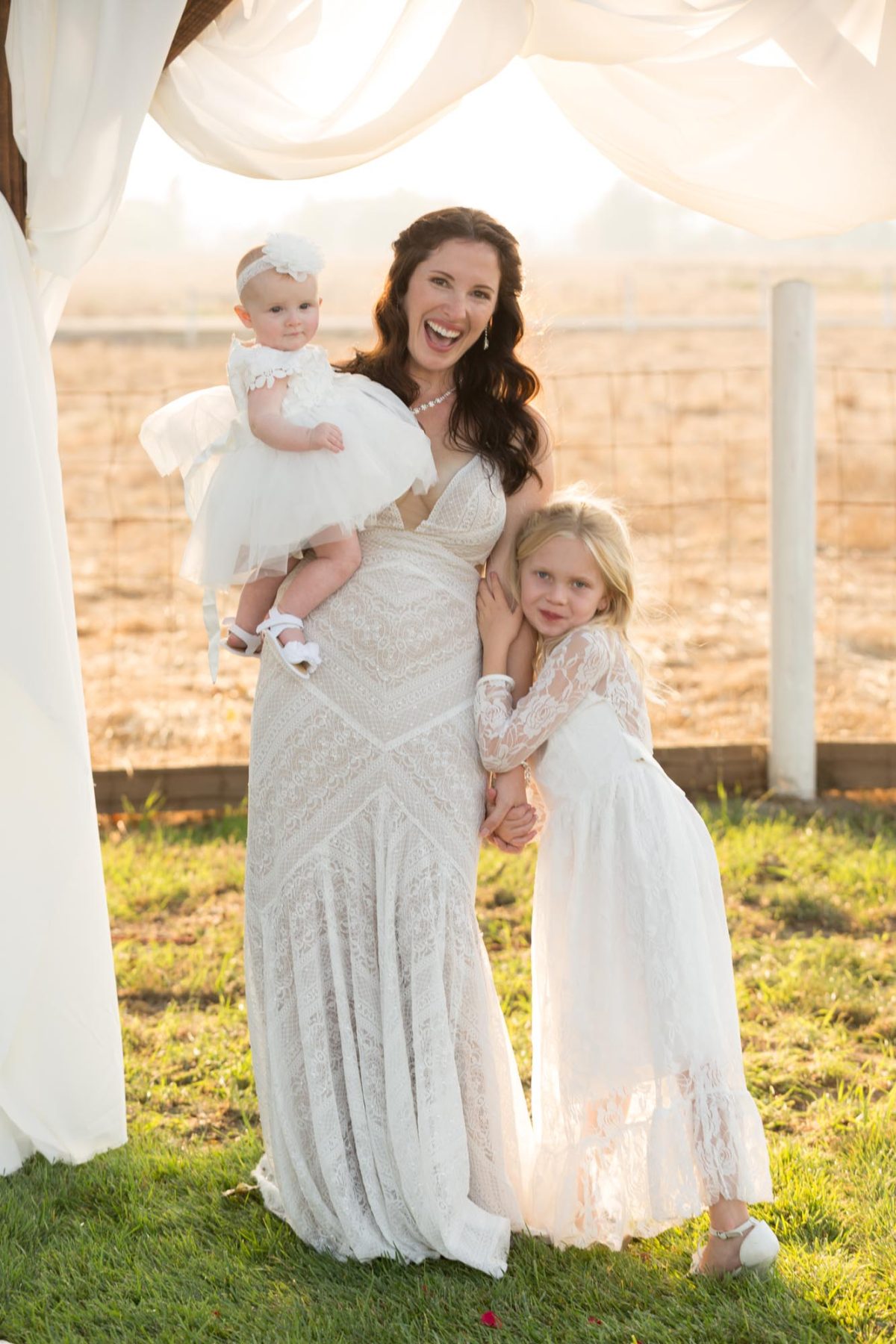 A mother beams with a wide smile while holding her baby daughter in a fluffy white tutu dress and standing beside her older daughter. This photo is a perfect example of family photos with mom and daughters. Warm golden hour light filters through flowing white drapes above them on a grassy field with soft backlighting highlighting their matching white lace outfits.