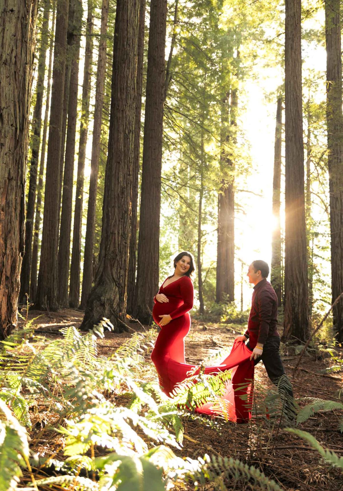 A pregnant woman in a long red dress smiles at her husband with both hands on her belly during their husband and wife maternity photos in a redwood forest. He holds the flowing train of her dress as bright golden sunlight streams through the tall trees creating beautiful lens flare.