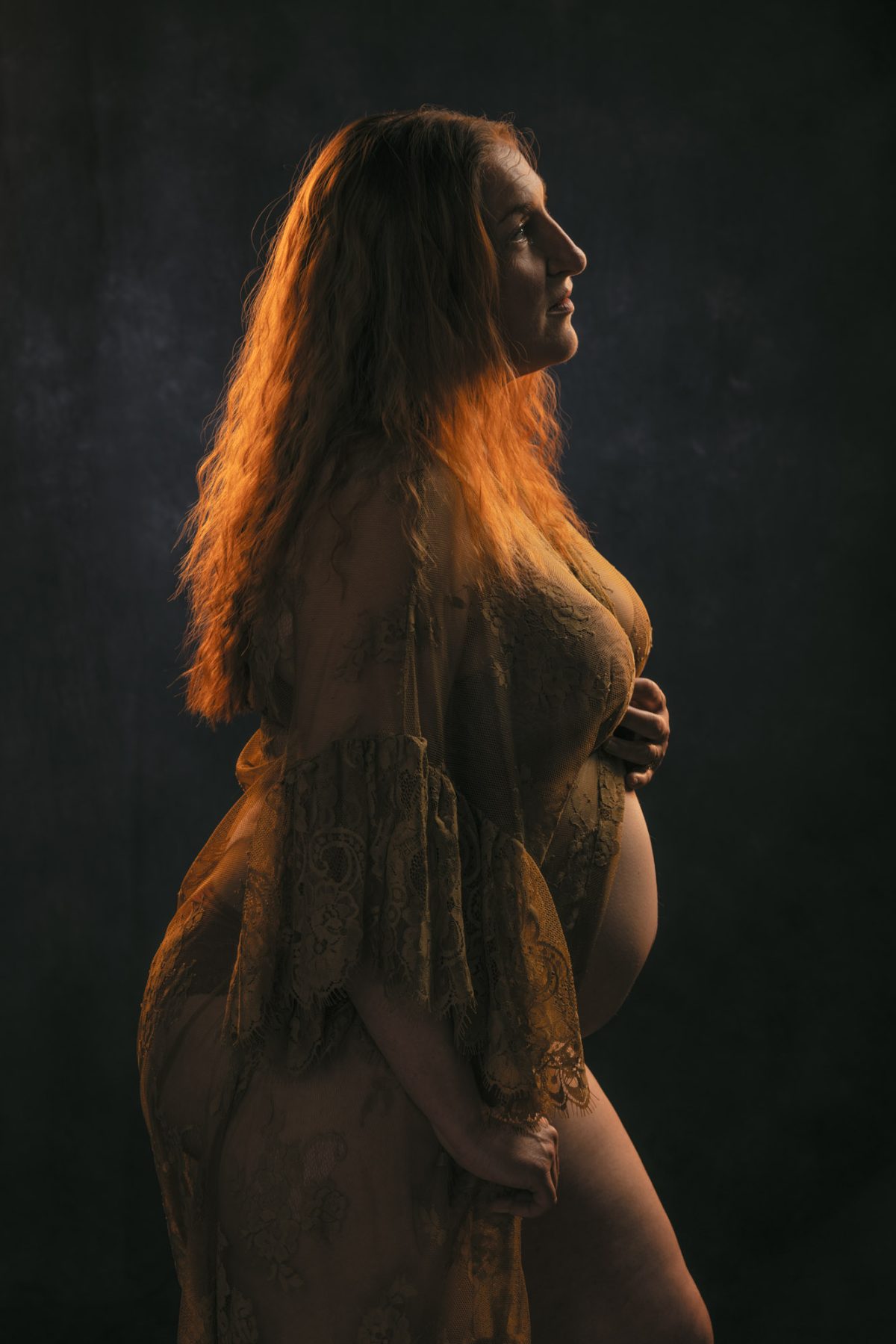 A pregnant woman with long flowing red hair stands in graceful profile while gently cradling her belly in warm dramatic lighting. This image was taken by maternity photographer Jason Guy as she wears a delicate lace robe during on location studio style session in Sonoma County.
