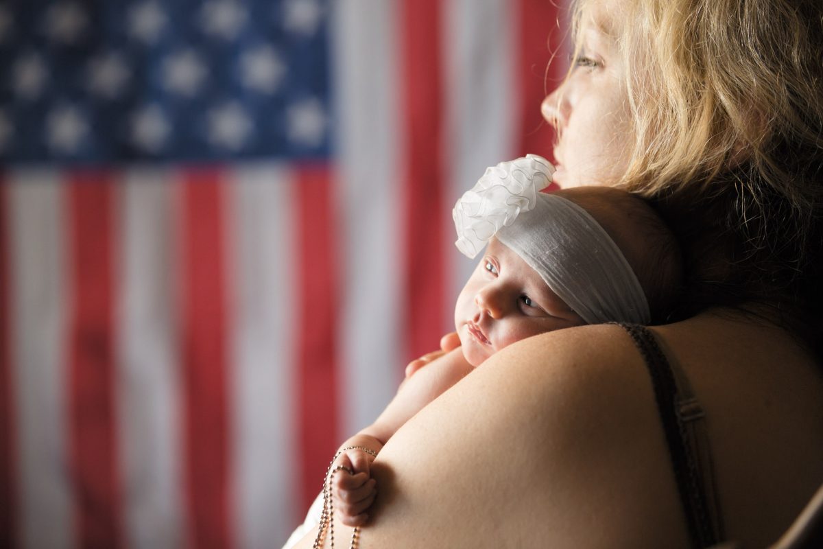 A military mother holds her baby daughter close during their military mother daughter portrait. She gazes to the side in profile as the infant rests on her shoulder wearing a large white ruffled headband while an American flag appears softly in the background.