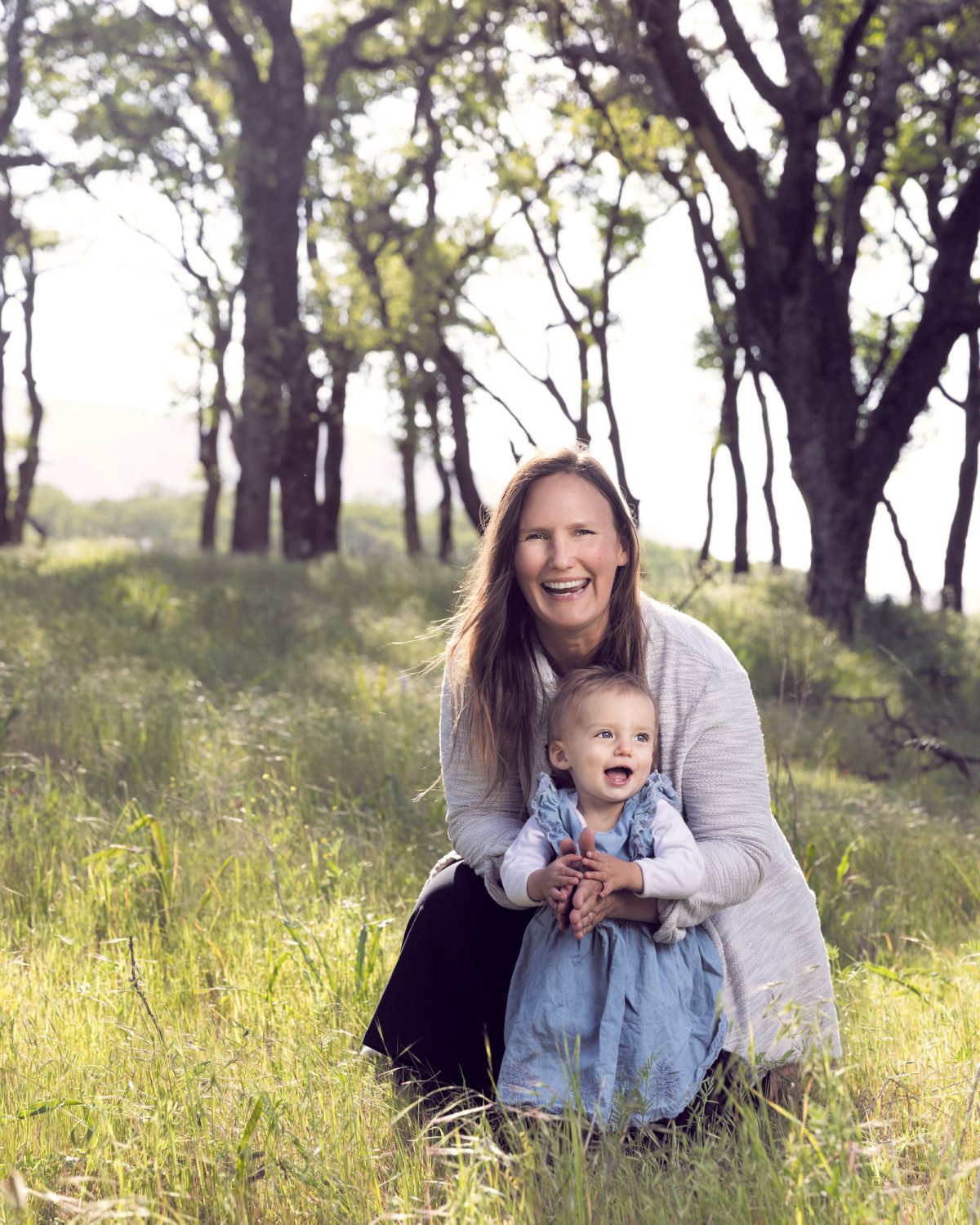 A smiling woman with long brown hair sits in a grassy field holding her baby daughter during mom and baby daughter photo session. Warm golden light illuminates the pair as the woman in a light gray cardigan and the baby in a blue dress both look happily at the camera against a backdrop of tall oak trees.