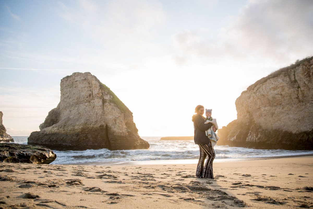 A mother holds her young son securely in her arms on a sandy beach during mother and son family photos. Warm golden sunlight bathes the pair as they stand before dramatic rock formations and rolling ocean waves at golden hour.