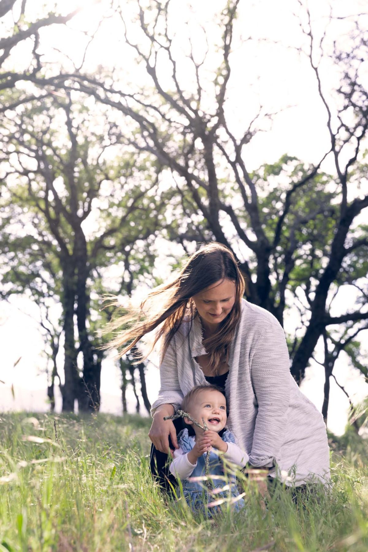 A woman with long brown hair blowing in the breeze crouches beside her young daughter for their mother daughter portraits in a sunlit grassy field. Bright sunlight highlights the lush green grass and background trees as the smiling child in a blue outfit holds a small branch and the mother in a light gray cardigan looks down at her tenderly.