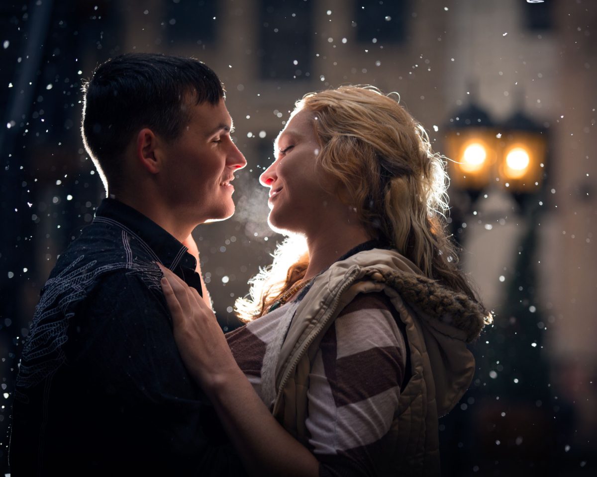 A young couple smiles lovingly at each other with snow gently falling around them during an engagement photo shoot. The woman rests her hand affectionately on the man's shoulder while warm golden light illuminates their faces from behind.