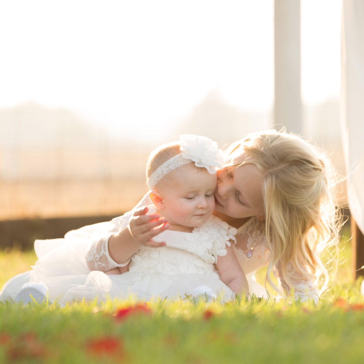 In this tender portrait of kid sisters an older blonde girl gently kisses her baby sister on the cheek as they lie together on a grassy lawn. Warm golden hour light creates a beautiful glow around them while both wear delicate white lace dresses.