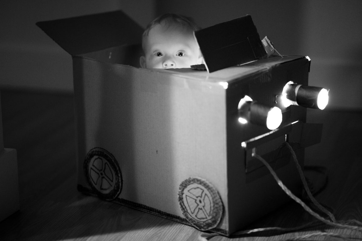 A wide-eyed baby peeks out from inside a handmade cardboard box car during a professional kids photo shoot. The black and white photograph features bright illuminated headlights glowing from the front of the creative vehicle with illustrated wheels along its side. The child gazes directly at the camera while nestled in the prop positioned on a wooden floor.