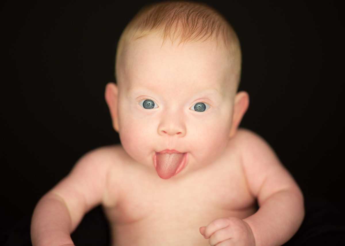 Bare chested infant shows tongue playfully during six month infant portrait. Soft illumination highlights bright blue eyes rounded face and light hair against black backdrop.