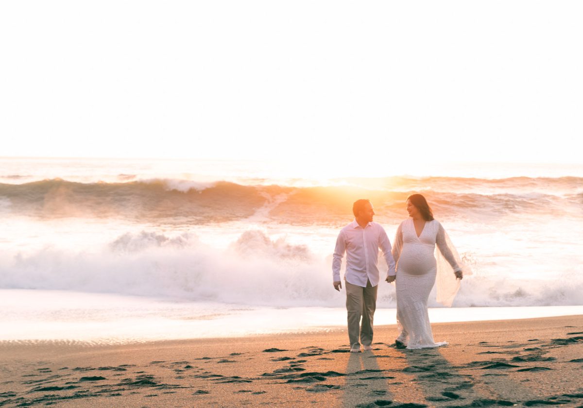 A pregnant woman in a white lace dress walks hand in hand with her husband along the beach during their Sonoma County beach couples photos. They smile at each other as golden sunset light illuminates the crashing ocean waves in the background.