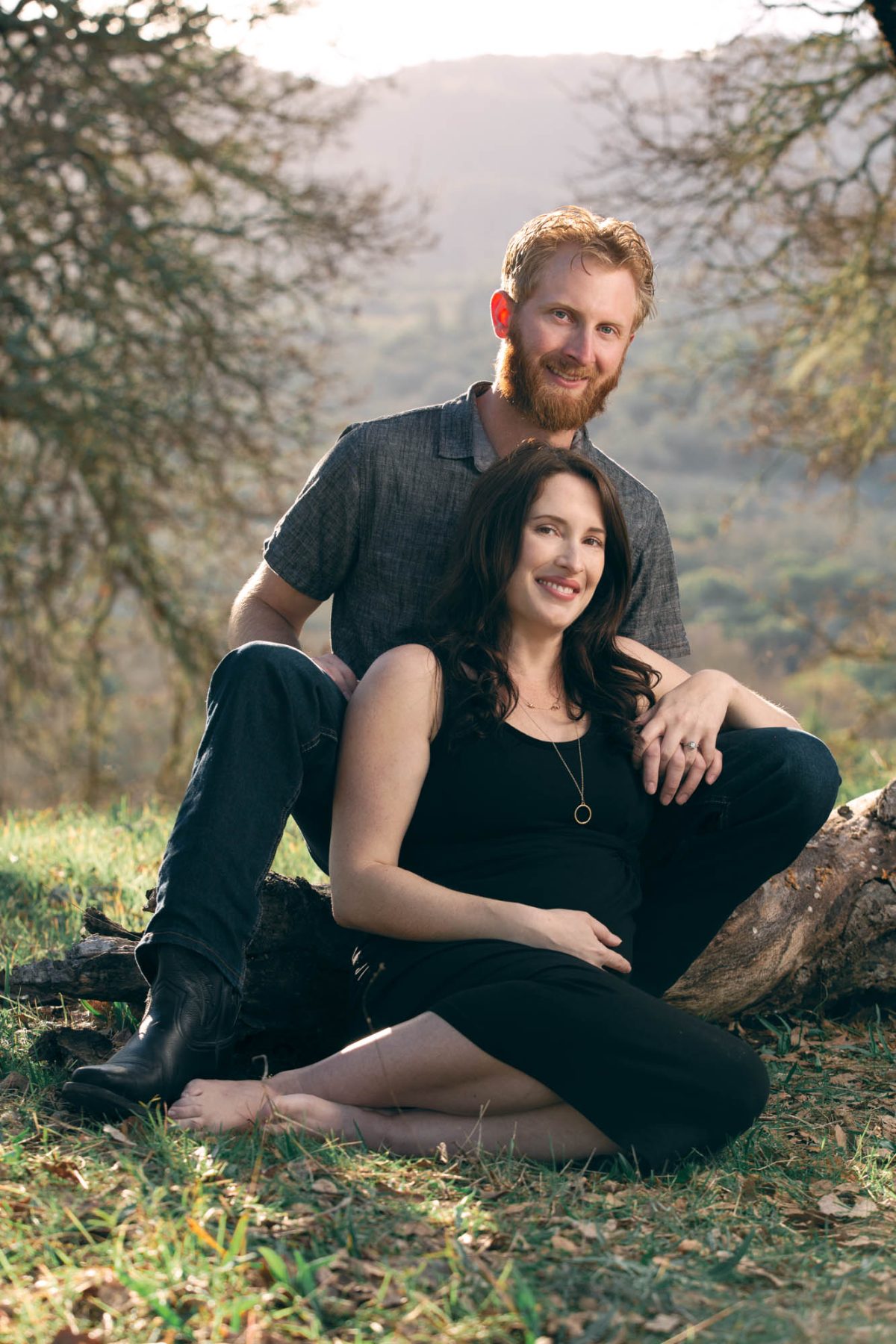 A smiling couple sits together on a fallen log during Sonoma Valley engagement photo session. The woman with long dark hair rests her hand gently on her belly while the man with red hair and beard places his arm around her in warm golden light against a hilly backdrop.