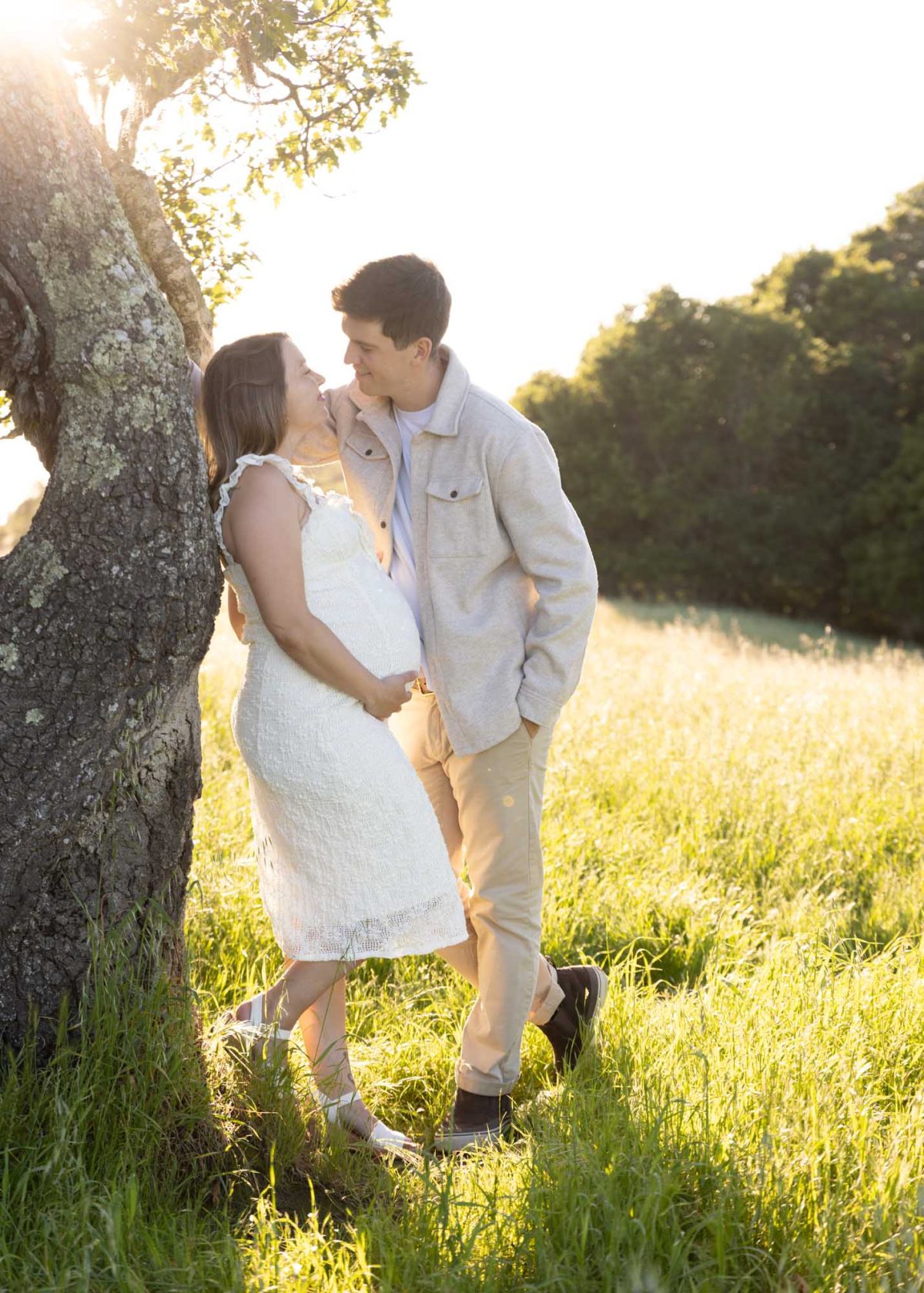 A pregnant woman in a white dress leans against a large tree with her husband during their recent Helen Putnam Regional Park couples photo session. She rests her hand on her belly while they smile at each other in the golden sunset light of the grassy field.