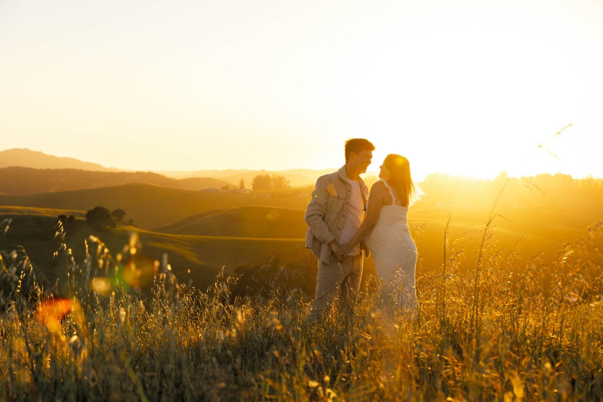 A pregnant woman and her partner hold hands and gaze at each other during their Helen Putnum couples photos. They stand close together in tall grass on a hillside as warm golden sunset light bathes the rolling hills behind them. She wears a white dress and he wears a light jacket with pants.