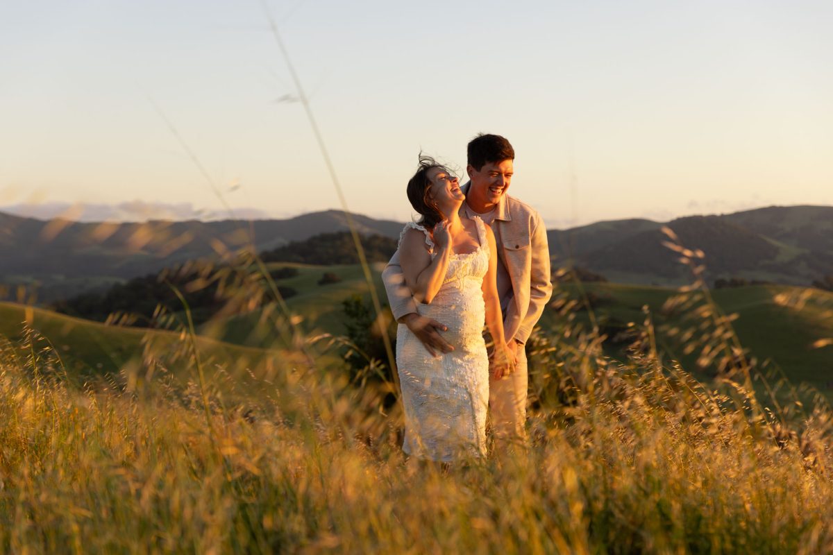 A pregnant woman and her partner share a joyful moment during their natural couples portraits in Petaluma. She laughs with her head tilted back and one hand near her neck while he smiles down at her with his arm around her waist and their hands clasped. Both stand close together in tall golden grass on a hillside as warm sunset light bathes the rolling hills behind them. She wears a white lace dress and he wears a light jacket with pants.